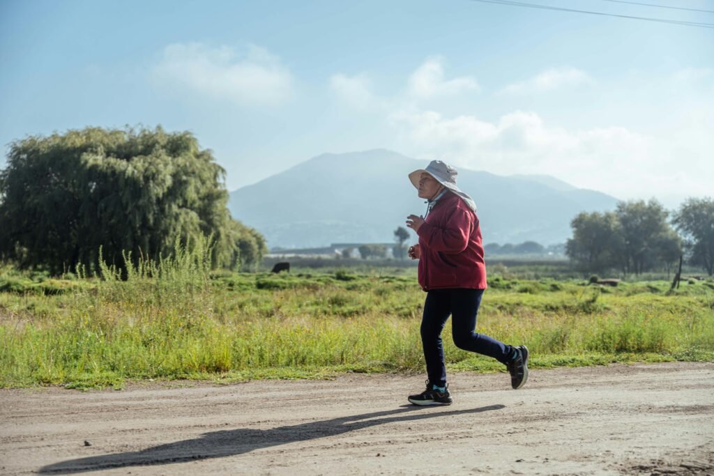 Senior woman jogging on a dirt road in a rural landscape with mountains in the background.
