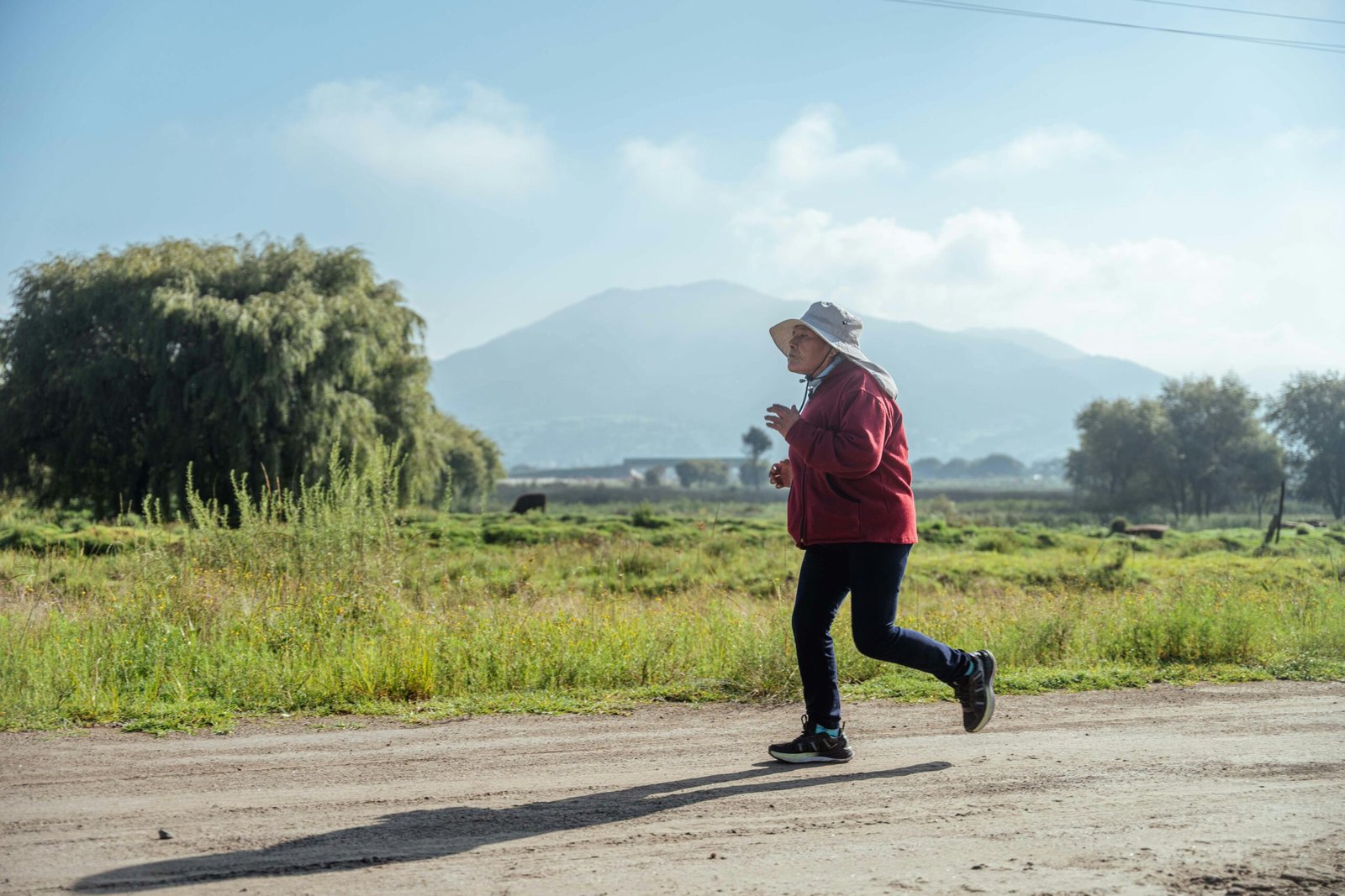 Senior woman jogging on a dirt road in a rural landscape with mountains in the background.
